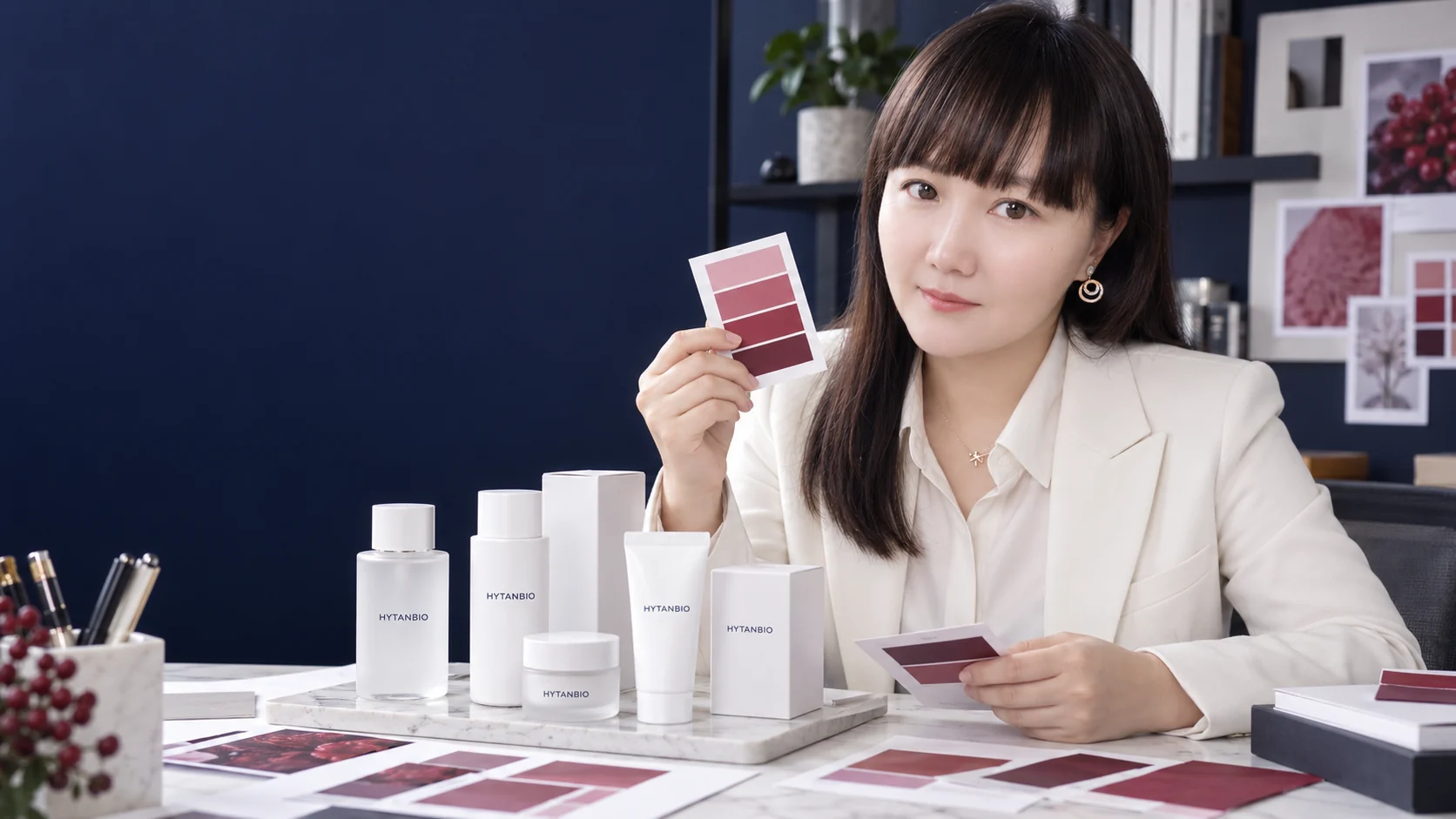 An East Asian woman designer with berry color swatches and unbranded skincare carton prototypes — illustrative development scene