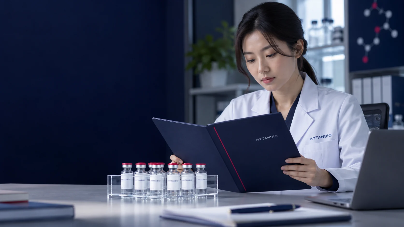 A closed research documentation folder and a clear vial rack beside a laptop in a quiet lab office — illustrative development scene