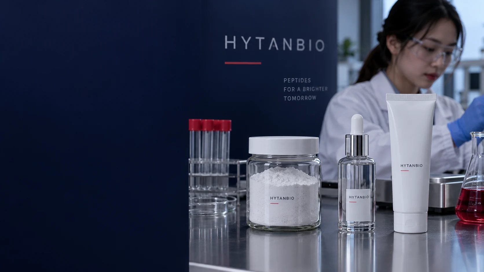 Macro environmental photograph of fine white powder inside one straight wide-neck capped glass jar beside an empty clear serum bottle and capped white squeeze tube, all standing on a dark metal formulation bench — illustrative development scene