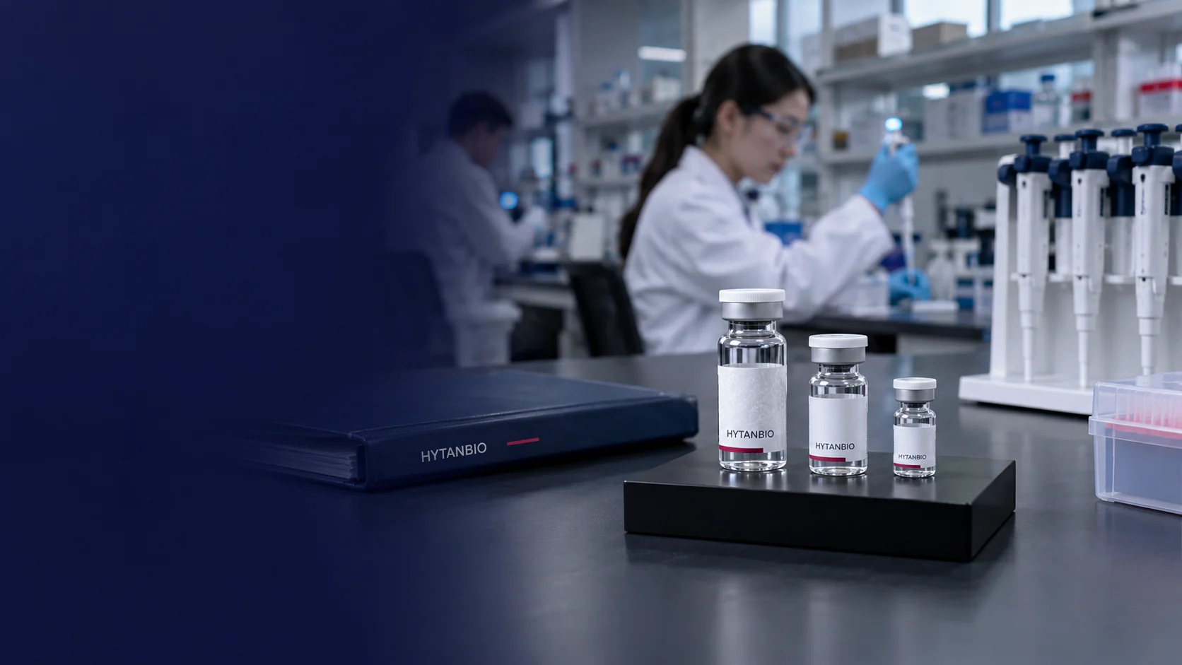 Three differently sized capped straight sample vials on a black square stand with a closed navy project folder and a capped pipette rack, photographed near bench level with far-off laboratory shelves — illustrative development scene