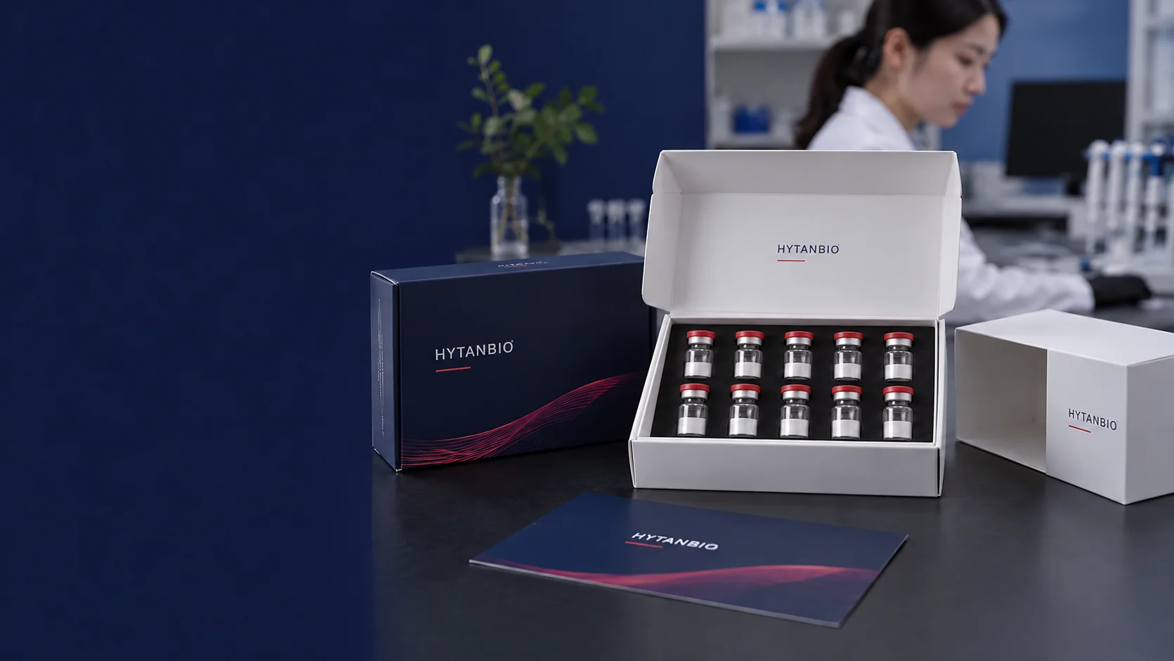 An open white product carton containing upright sealed lyophilized vials in a foam insert, a separate empty carton sleeve and navy packaging proof lying on a charcoal table, no hands — illustrative development scene