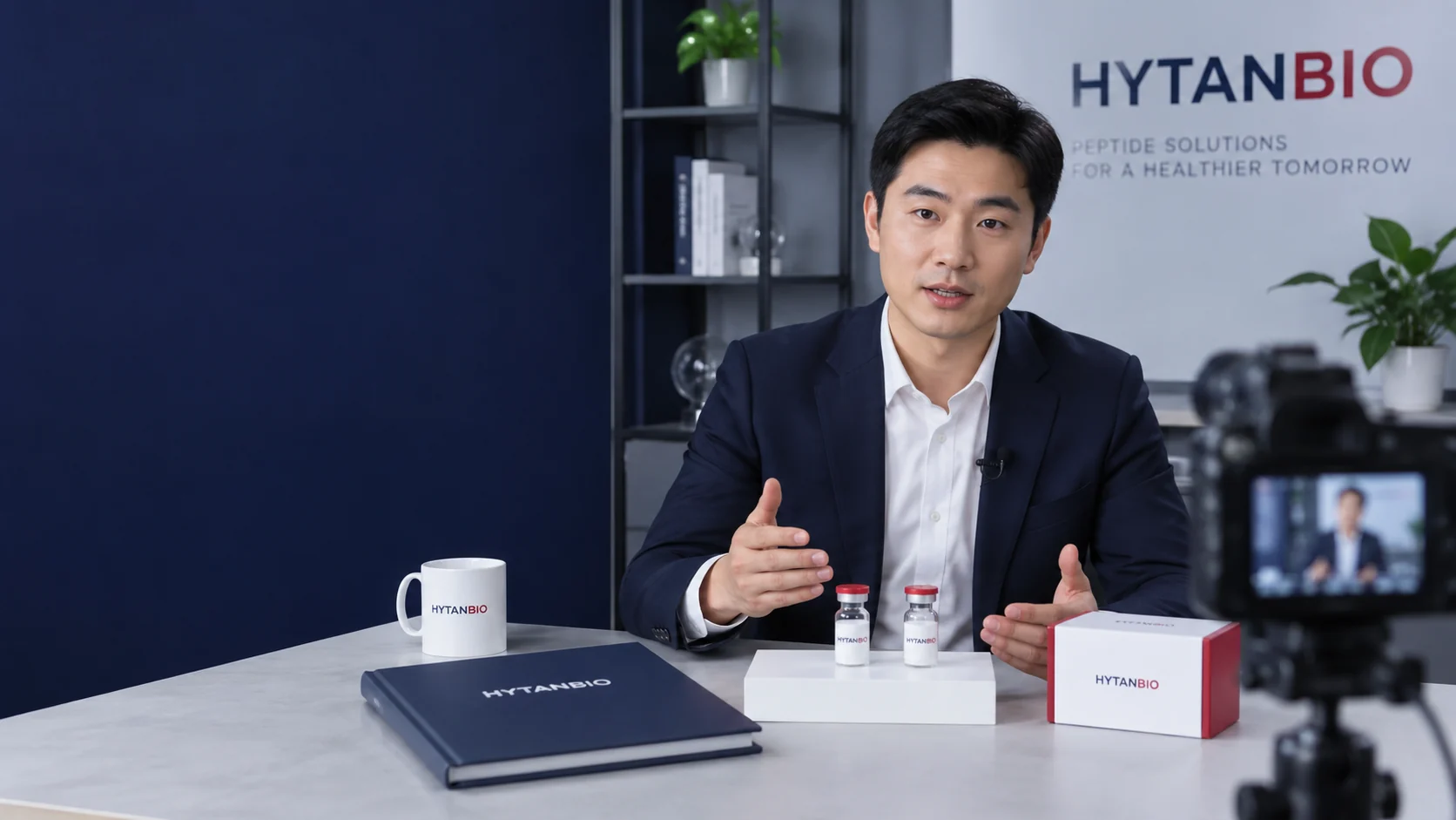 An East Asian male product specialist recording a lyophilized supply brief, with two capped vials standing on a white rectangular stand and a closed navy binder beside a berry-edged carton — illustrative development scene