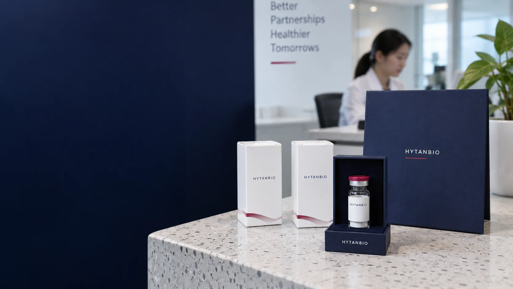 An individually boxed sealed lyophilized sample vial standing vertically within a tabletop display cradle, two unopened white sample cartons and a plain navy presentation folder on a pale terrazzo dispatch counter — illustrative development scene