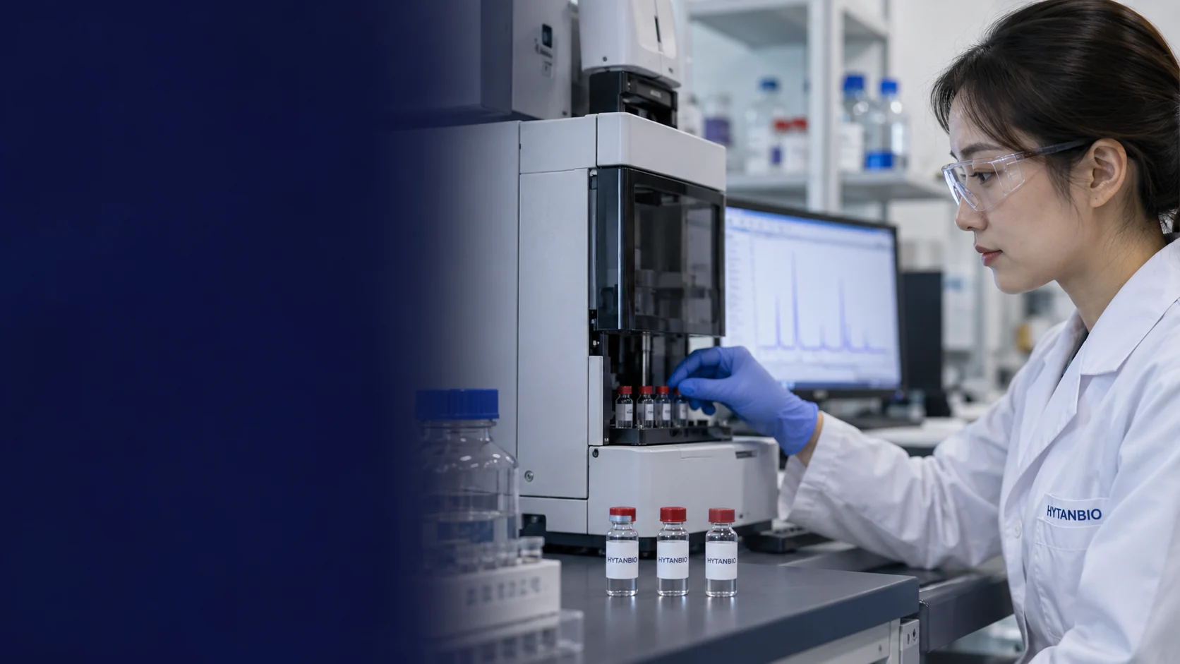 A desktop instrument with small enclosed sample tray, three sealed research vials on a metal platform and blurred unreadable chromatogram on a distant monitor, ordinary laboratory setting — illustrative development scene