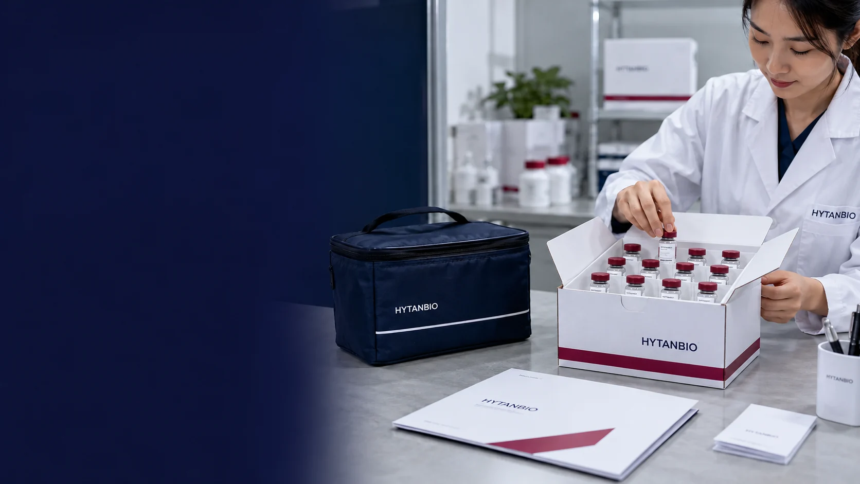 Sealed small vials packed upright inside a white shipping insert next to a closed navy thermal pouch and flat paperwork folder; a muted berry stripe on the carton, pale concrete surface — illustrative development scene