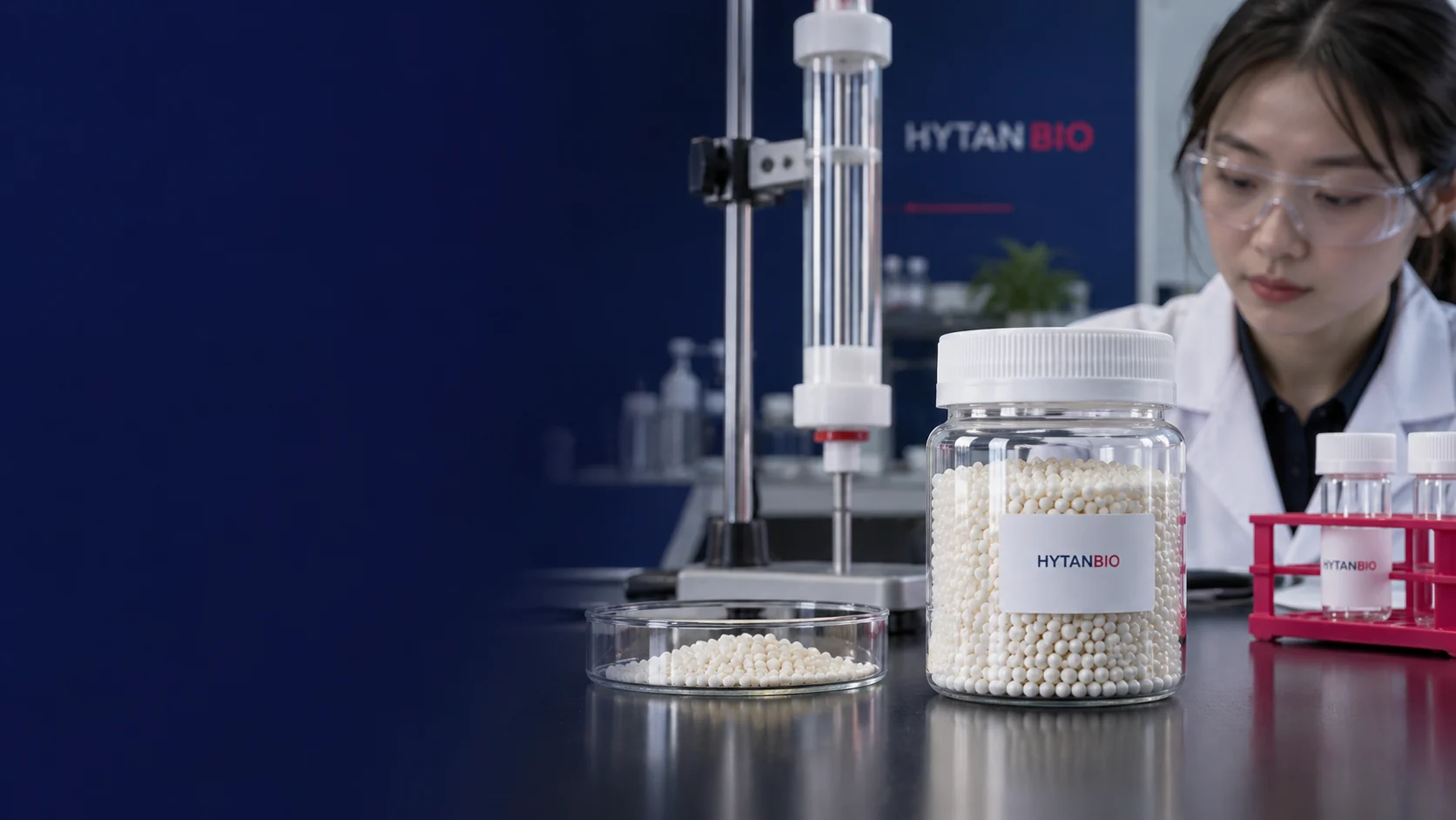 Macro photograph of a capped transparent wide-mouth jar containing tiny cream-colored resin beads, a dry clear sample dish beside it and a clean empty glass column in the background, no experiment shown — illustrative development scene