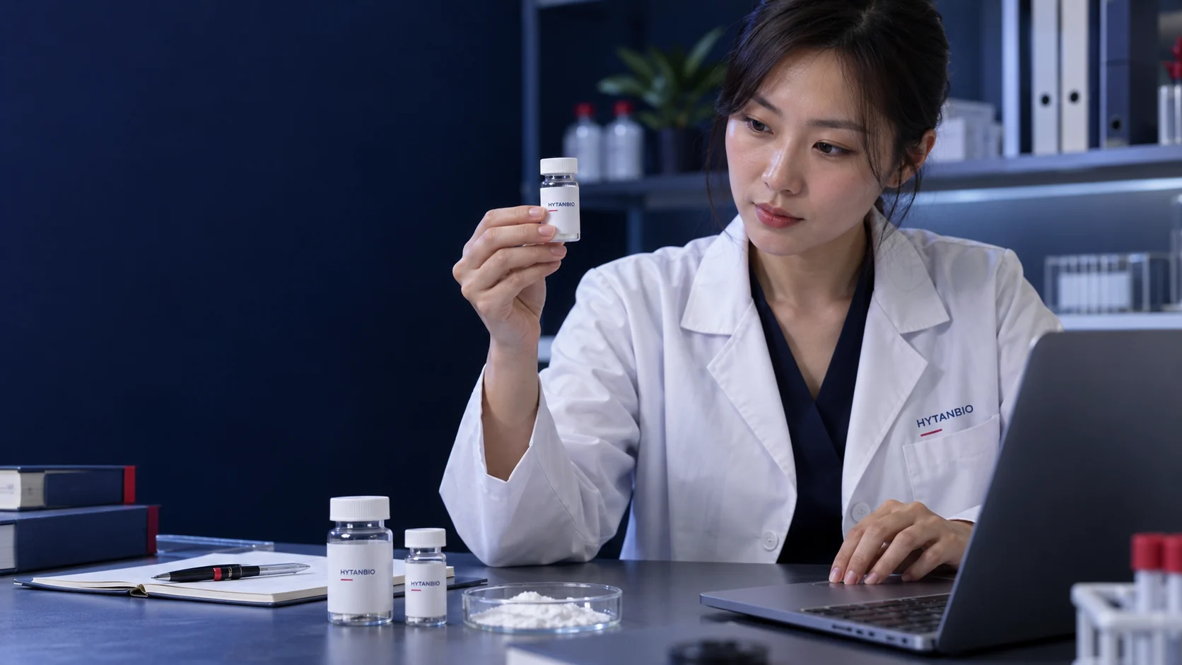 An East Asian technical colleague compares a material sample bottle with a catalog on a laptop, no legible fabricated data — illustrative development scene