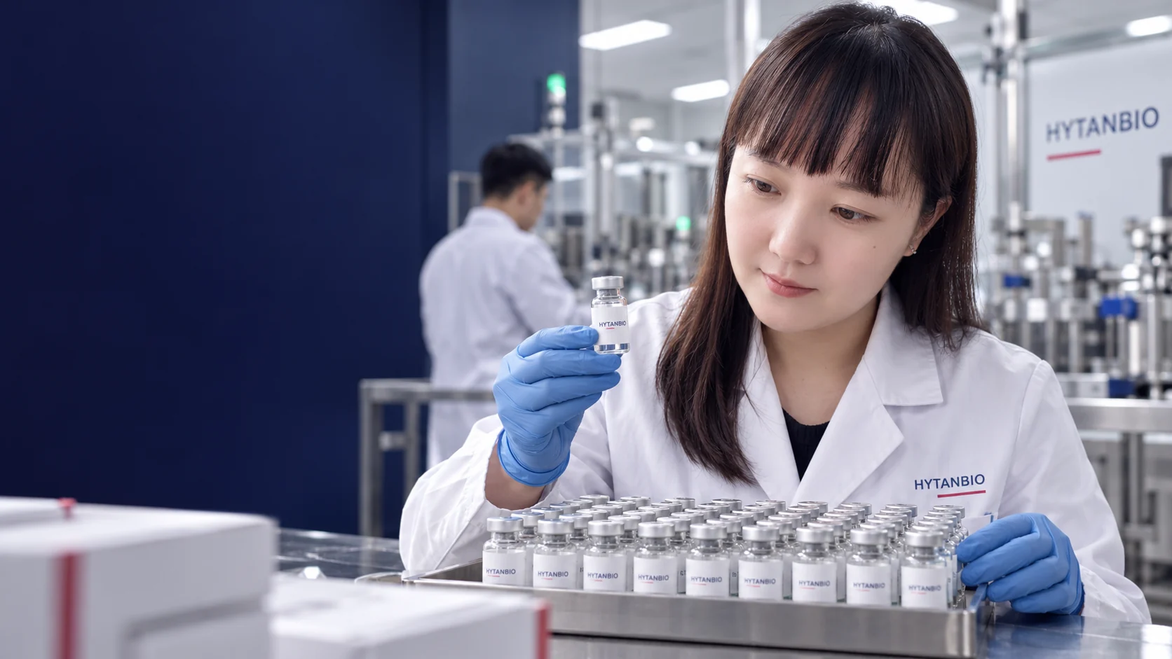 An East Asian manufacturing coordinator reviews a sealed vial tray in a bright workshop — illustrative development scene