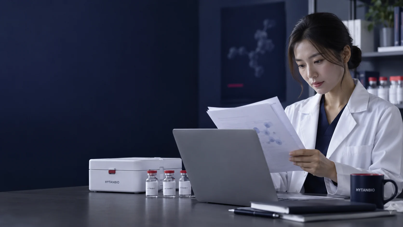 An East Asian specialist reads out-of-focus technical pages beside a laptop and closed sample case — illustrative development scene