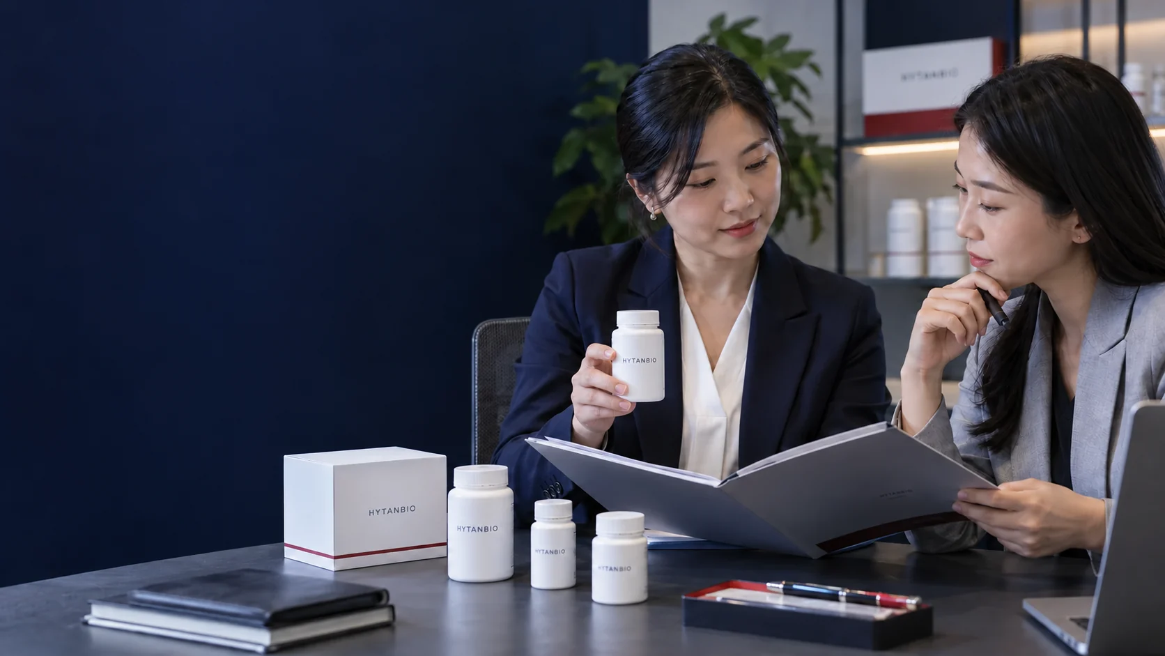 Two East Asian project colleagues compare a specification folder and a neutral packaging prototype at a meeting table — illustrative development scene