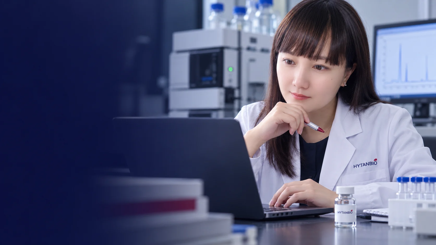 An East Asian analyst studies a softly blurred chromatogram at a laptop with a sealed sample nearby — illustrative development scene