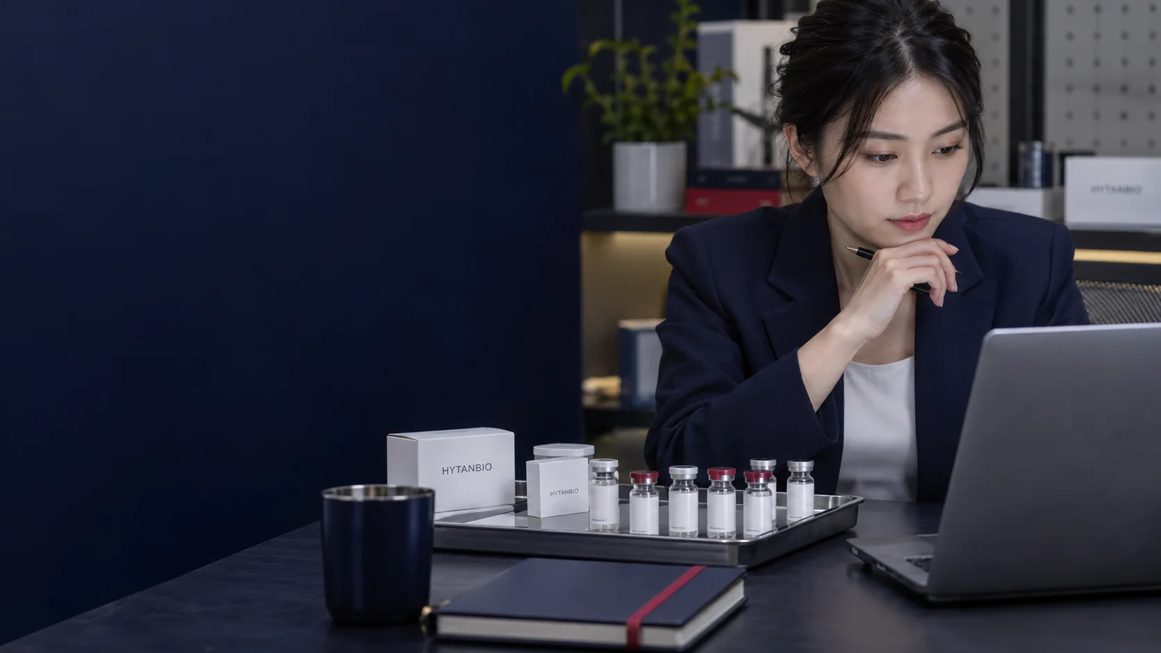 An East Asian coordinator studies a laptop beside a packaging reference tray and a notebook — illustrative development scene