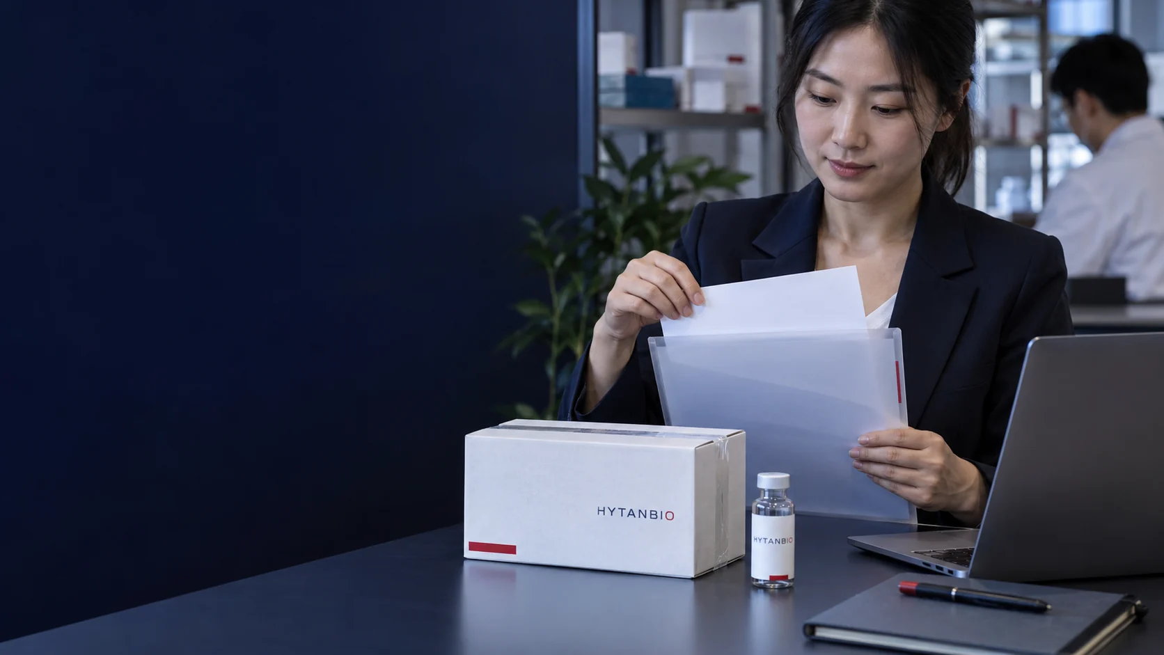 A sealed sample carton, paper document sleeve and laptop showing no legible private information — illustrative development scene